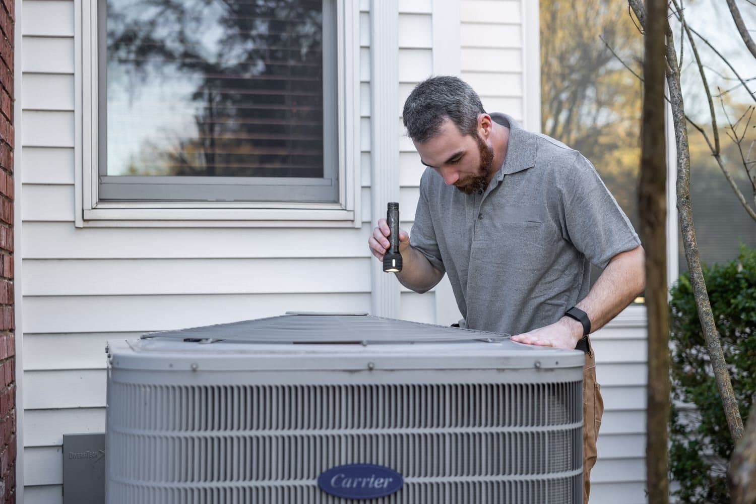 Residential HVAC technician inspecting an outdoor condenser beside a home